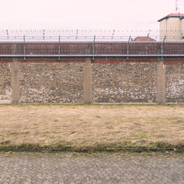 Die Außenmauer der JVA Rennelberg. Eine hohe Mauer aus Backstein und Stacheldraht umgibt eine freie Wiesenfläche.